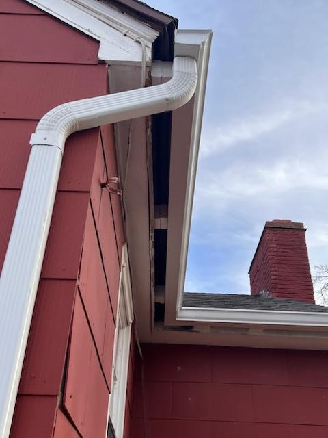 Close-up of white 5-inch residential gutters and downspout installed on a red house to direct rainwater away from the roof and foundation.
